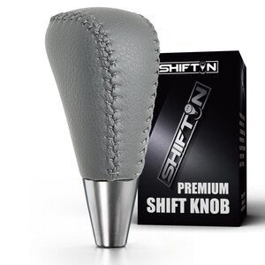 May include: A grey leather gear shift knob with visible stitching and a metallic silver base. The product is displayed next to a black box featuring the text SHIFTON and PREMIUM SHIFT KNOB in white lettering, with a small gear icon in the logo.