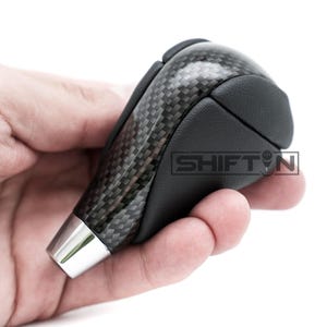 May include: A carbon fiber shift knob with a black leather grip. The shift knob has a silver base and the text "SHIFT N" is visible on the side. The carbon fiber has a woven pattern.