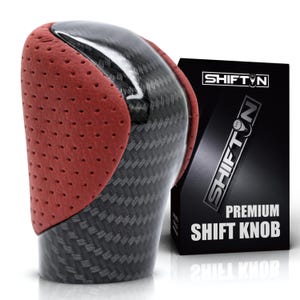 May include: A gear shift knob featuring a black carbon fiber pattern and perforated red side panels. Next to it stands a black rectangular box with the text SHIFTN and PREMIUM SHIFT KNOB printed in white, along with a logo graphic of a shift knob.