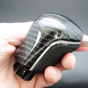 May include: A carbon fiber shift knob with a glossy finish. The knob has a textured pattern and a black, perforated leather grip. The text "SHIFTIN" is visible on the side. The shift knob is held in a hand.
