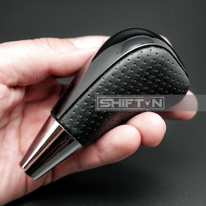 May include: A black and silver car shift knob with the text "SHIFT N". The top of the knob is glossy black, and the sides are black perforated leather. The base is a shiny silver color.