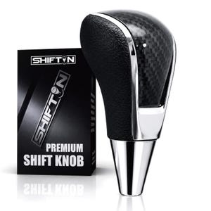 May include: A gear shift knob featuring a black textured grip, a carbon fiber pattern insert, and a polished silver metallic base. Next to it stands a black rectangular box with the text SHIFTN, PREMIUM SHIFT KNOB, and a gear icon printed on it.