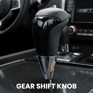 May include: A black and chrome gear shift knob with the text "GEAR SHIFT KNOB" at the bottom. The top of the knob is black leather, and the bottom is chrome. The knob is in a car interior.