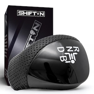 May include: A black automotive gear shift knob featuring a perforated texture on the sides and a smooth, glossy top surface. The top displays a white gear pattern with the letters R, N, D, and B, accompanied by directional arrows. The product is shown in front of its black branded SHIFTIN packaging box.