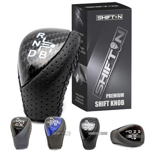 May include: A black leather shift knob with a carbon fiber pattern and white lettering that reads "R N D B". The knob is surrounded by four other shift knobs in different colors, including black, blue, and gray. The image also includes a box with the text "SHIFT'N PREMIUM SHIFT KNOB".