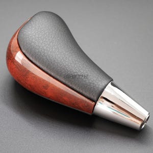 May include: A car gear shift knob with a black leather top and a polished wood grain base. The bottom portion is chrome. The word "SHIFTIN" is printed on the leather.