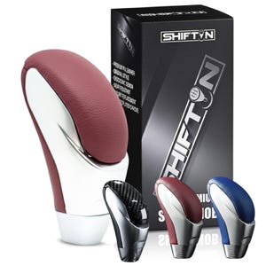 May include: A collection of car shift knobs in various colors, including red, blue, and carbon fiber. The main knob is a burgundy and white design with a chrome base. The box features the "SHIFT N" logo.
