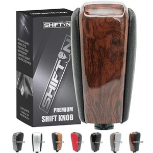 May include: A wood grain shift knob with a black leather base. The knob is a replacement part for a car. The knob is shown in a box with the brand name "SHIFTIN" on it. The box also shows other colors and styles of shift knobs.