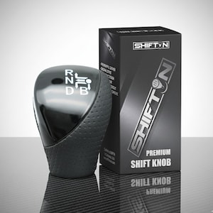 May include: A black shift knob with a glossy top and a textured grip. The knob has white lettering indicating gear positions: R, N, D, and B. A black box with the text "SHIFT N" and "PREMIUM SHIFT KNOB" is next to the knob.