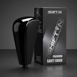 May include: A black premium shift knob with a glossy top and textured side panel, next to a black box with the "SHIFT N" logo and "PREMIUM SHIFT KNOB" text. The shift knob is designed for a vehicle's gear shift.