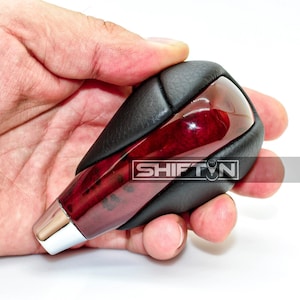 May include: A car shift knob with a red wood grain design encased in a black leather grip. The base is chrome, and the text "SHIFT N" is visible on the side.