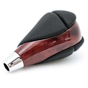 May include: A car gear shift knob with a polished wood grain finish and black leather accents. The shift knob has a chrome-colored base and the text "SHIFTYN" is visible on the wood grain.