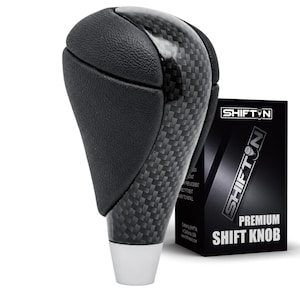 May include: A black gear shift knob featuring a textured leather-like grip and a glossy carbon fiber center inlay. The base is a smooth silver-toned metal. Beside it stands a black product box labeled SHIFTON PREMIUM SHIFT KNOB in white text.