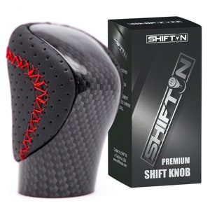 May include: Black leather and carbon fiber shift knob with red stitching. The knob is packaged in a black box with the brand name "SHIFT'N" printed on it. The box also says "Premium Shift Knob".