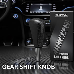 May include: A carbon fiber and black leather gear shift knob with a silver base. The product is displayed in a car interior with the text "SHIFT N" and "PREMIUM SHIFT KNOB" on the packaging. The words "GEAR SHIFT KNOB" are also visible.