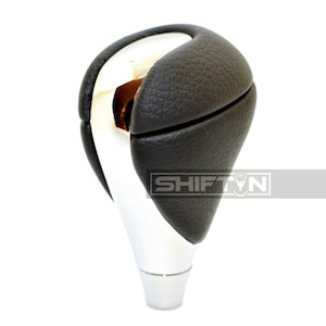 May include: A car gear shift knob with a black leather top and a silver base. The top is split into two sections. The text "SHIFT" is visible on the side of the knob.