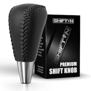 May include: A black leather gear shift knob with visible stitching and a silver metallic base, positioned next to a black rectangular product box labeled SHIFTIN PREMIUM SHIFT KNOB with the brand logo and additional text printed on the side.