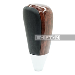 May include: A brown wood and black leather gear shift knob with a silver base. The knob has a wood grain pattern and the leather is smooth. The base is chrome.