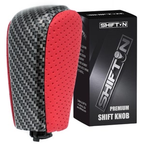 May include: A red and black carbon fiber shift knob with a perforated leather top. The knob is designed to replace the factory shift knob in a car. The packaging says "SHIFT'N PREMIUM SHIFT KNOB".
