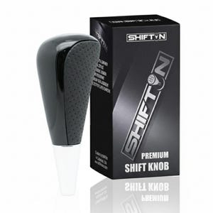 May include: A black shift knob with a textured grip and a white base, next to a black box. The box features the text "SHIFT N" in silver and "PREMIUM SHIFT KNOB". The shift knob is designed for a vehicle's gear shift.