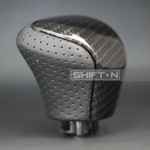 May include: A black and gray shift knob with a carbon fiber weave pattern. The top portion is a glossy black, while the lower section features a perforated leather texture. The text "SHIFT N" is visible on the side.
