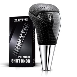 May include: A black gear shift knob featuring a perforated texture on the top and a carbon fiber pattern on the side, paired with a polished silver metallic base. Next to it stands a black box labeled SHIFTN PREMIUM SHIFT KNOB in white text.