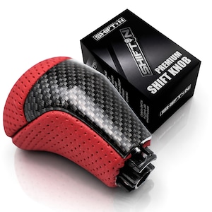 May include: A gear shift knob featuring a black carbon fiber center panel and red perforated side panels. The knob is positioned in front of a black rectangular box labeled SHIFTN PREMIUM SHIFT KNOB with white text and a logo on the top and side.