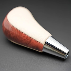 May include: A car gear shift knob with a cream-colored top and a wood-grain patterned base. The base is a reddish-brown color. The bottom of the knob is a shiny, silver metal.
