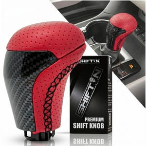 May include: A gear shift knob featuring a red perforated leather top and a black carbon fiber pattern base with black stitching. The product is displayed next to its black packaging box labeled SHIFTON PREMIUM SHIFT KNOB with a logo.