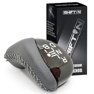 May include: A gray and black shift knob with a stitched leather grip and a glossy black top. The top has white lettering indicating gear positions. A black box with the "SHIFT N" logo is in the background.