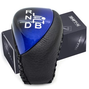 May include: A black and blue car gear shift knob with white lettering indicating the gear positions. The knob is partially wrapped in black leather and sits next to a black box with the word "SHIFTN" printed on it.