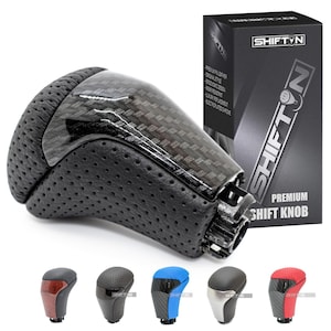 May include: Black and carbon fiber shift knob with a perforated leather grip. The product is displayed with its packaging, which features the brand name "SHIFT'N" and the words "Premium Shift Knob". Several other shift knobs in different colors are shown below.