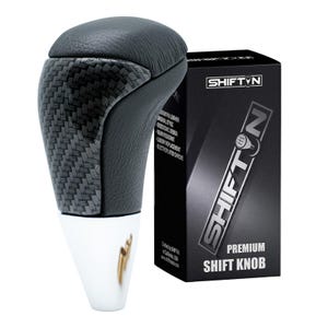 May include: A black and gray shift knob with a carbon fiber design and a leather top. The shift knob is next to a black box with the text "SHIFT N" and "PREMIUM SHIFT KNOB".