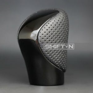 May include: A black car gear shift knob with a perforated leather grip and a glossy top. The word "SHIFT N" is printed on the side of the knob. The knob is designed for a vehicle's transmission.