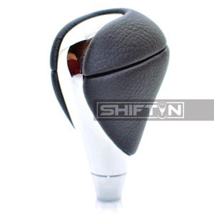 May include: A chrome and black shift knob with the text "SHIFTN". The top of the knob is covered in black textured leather. The base is a shiny chrome color.