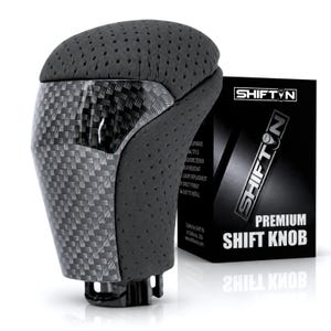May include: A black gear shift knob featuring a perforated texture on the top and sides, combined with a glossy carbon fiber pattern on the front. Next to it stands a black product box labeled SHIFTN PREMIUM SHIFT KNOB with white text.