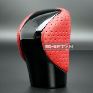 May include: A car shift knob with a perforated red leather grip and glossy black accents. The shift knob has the text "SHIFT N" on the side. The base of the knob is black.