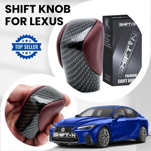 May include: A gear shift knob for a Lexus vehicle featuring a carbon fibre pattern centre panel flanked by dark red textured side panels. The product is displayed alongside its black retail box labelled SHIFTN and a blue Lexus saloon.