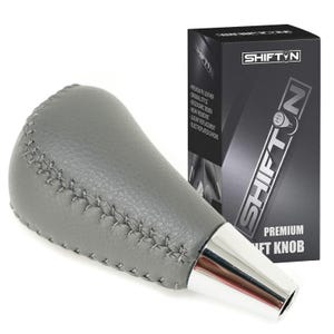 May include: A gray leather shift knob with silver accents and visible stitching. The shift knob is next to a black box with the text "SHIFT N" and "PREMIUM SHIFT KNOB".
