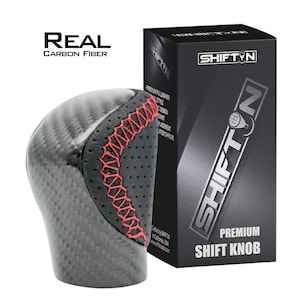 May include: A carbon fiber shift knob with a black leather grip and red stitching. The product is packaged in a black box with the text "SHIFT N" and "PREMIUM SHIFT KNOB". The text "REAL CARBON FIBER" is also visible.