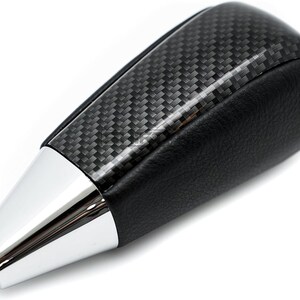 May include: A black and silver car gear shift knob. The top portion has a carbon fiber weave pattern, while the bottom is black leather. The tip is a shiny silver color.