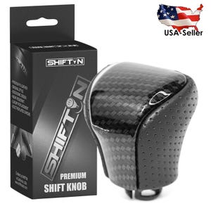 May include: A black and carbon fiber shift knob with a perforated leather grip. The product is packaged in a black box with the text "SHIFTN PREMIUM SHIFT KNOB". The box also includes product details.