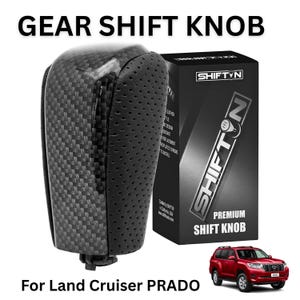 May include: A gear shift knob with a black carbon fiber pattern and perforated black side panels. The product is displayed next to its black packaging box labeled SHIFTN PREMIUM SHIFT KNOB and a small image of a red Land Cruiser PRADO.