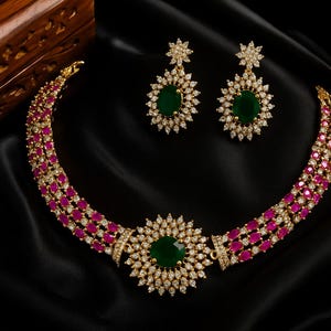 May include: A gold-coloured necklace and matching earrings set. The necklace features a large, round green stone at the centre, surrounded by smaller white stones. The necklace and earrings have a floral design with pink and white stones.
