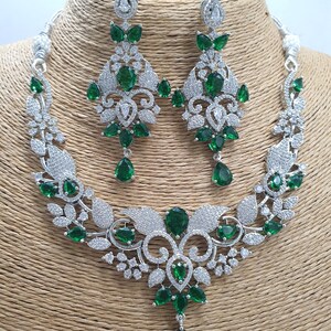May include: A silver necklace and earrings set with a floral design and green emerald-like stones. The necklace has a pendant with a teardrop-shaped stone.