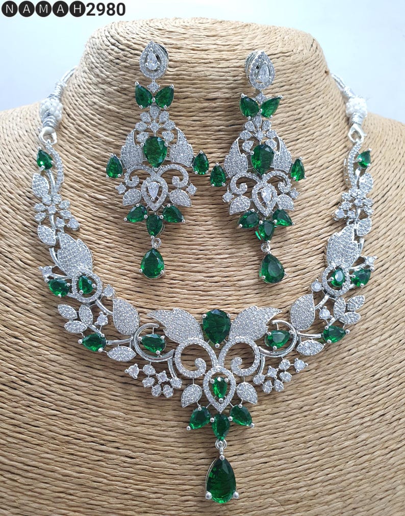 May include: A silver necklace and earrings set with a floral design and green emerald-like stones. The necklace has a pendant with a teardrop-shaped stone.