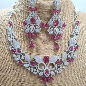 May include: A silver necklace and earrings set with a floral design and pink stones. The necklace has a pendant with a large pink stone in the center.