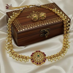May include: A gold-toned necklace and matching earrings set with red and green accents and small white pearls. The jewelry is displayed on a white satin surface next to a wooden jewelry box with gold detailing.