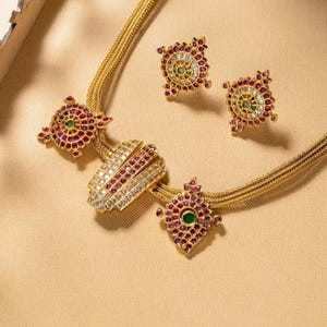 Puede incluir: Un collar de oro con un colgante y pendientes. El colgante es de forma rectangular con un diseño de piedras blancas y rojas. Los pendientes son redondos con un diseño de piedras blancas y rojas.