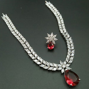 Puede incluir: Un collar de plata con un colgante de piedra roja en forma de lágrima y pendientes a juego. El collar y los pendientes están adornados con piedras de circonita cúbica transparentes.